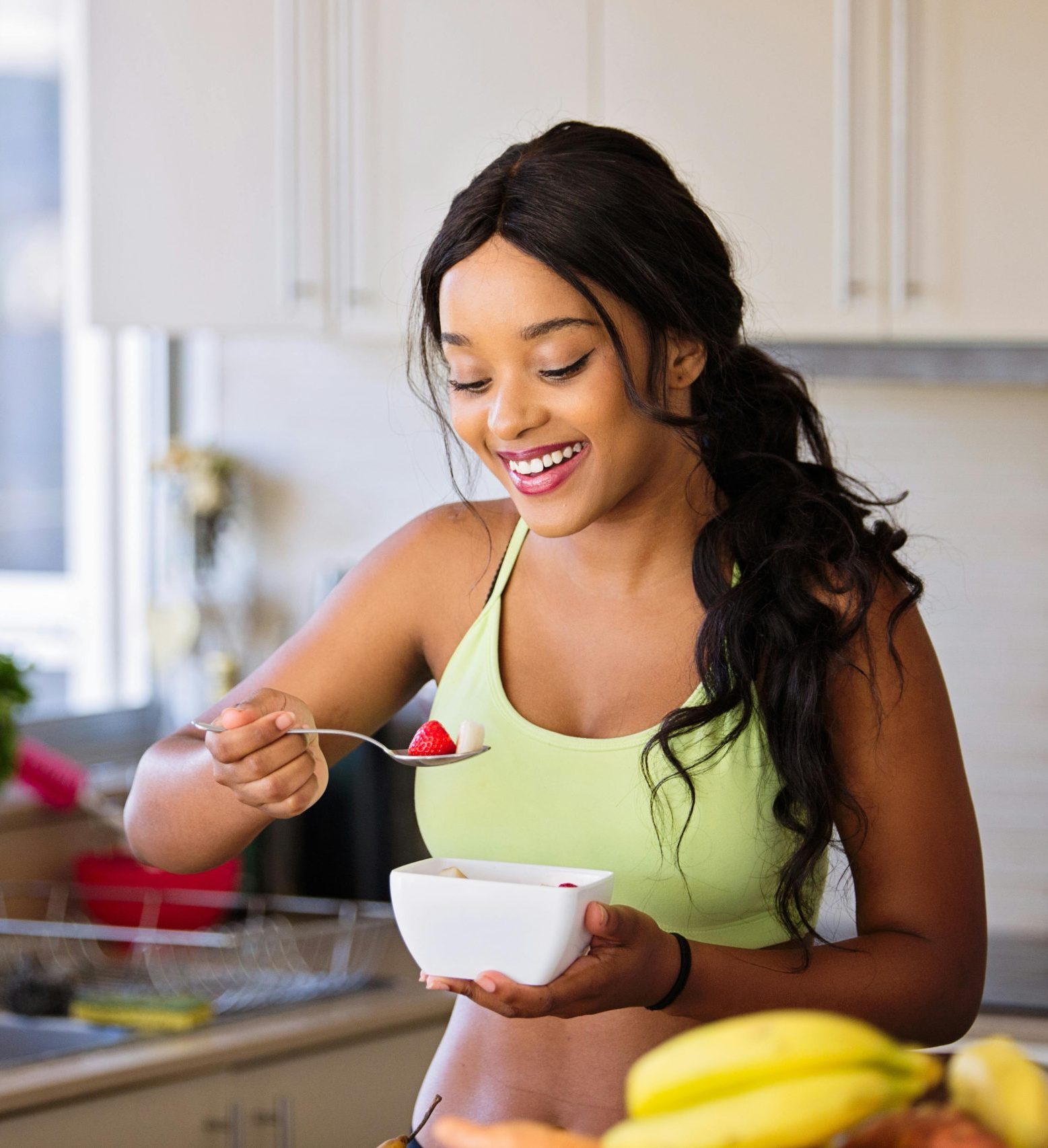 Smiling woman eating a nutritious fruit bowl in a bright kitchen setting.