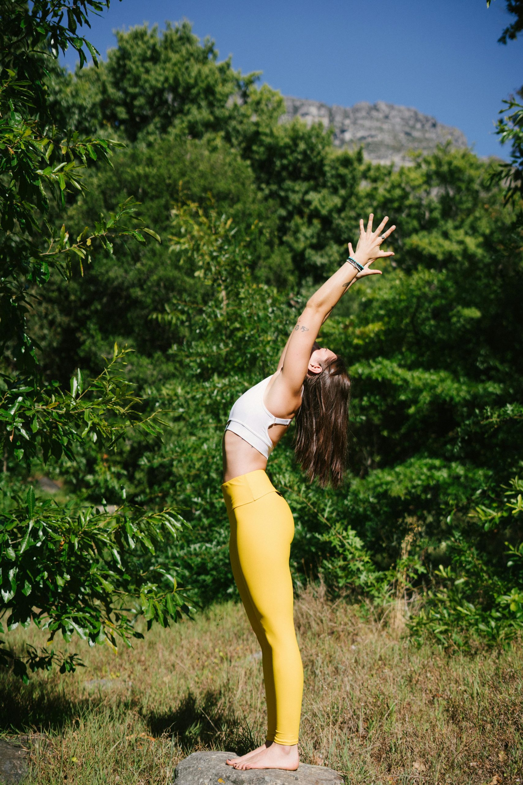 A woman in yellow leggings performs a yoga pose outdoors surrounded by green trees.